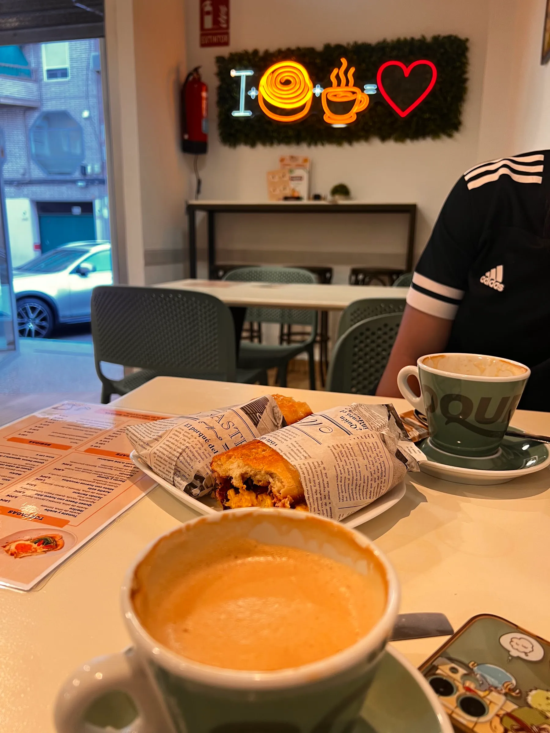 Una mesa con café con leche y un brioche envuelto en papel de periódico; detrás, las sillas verdes y el neón