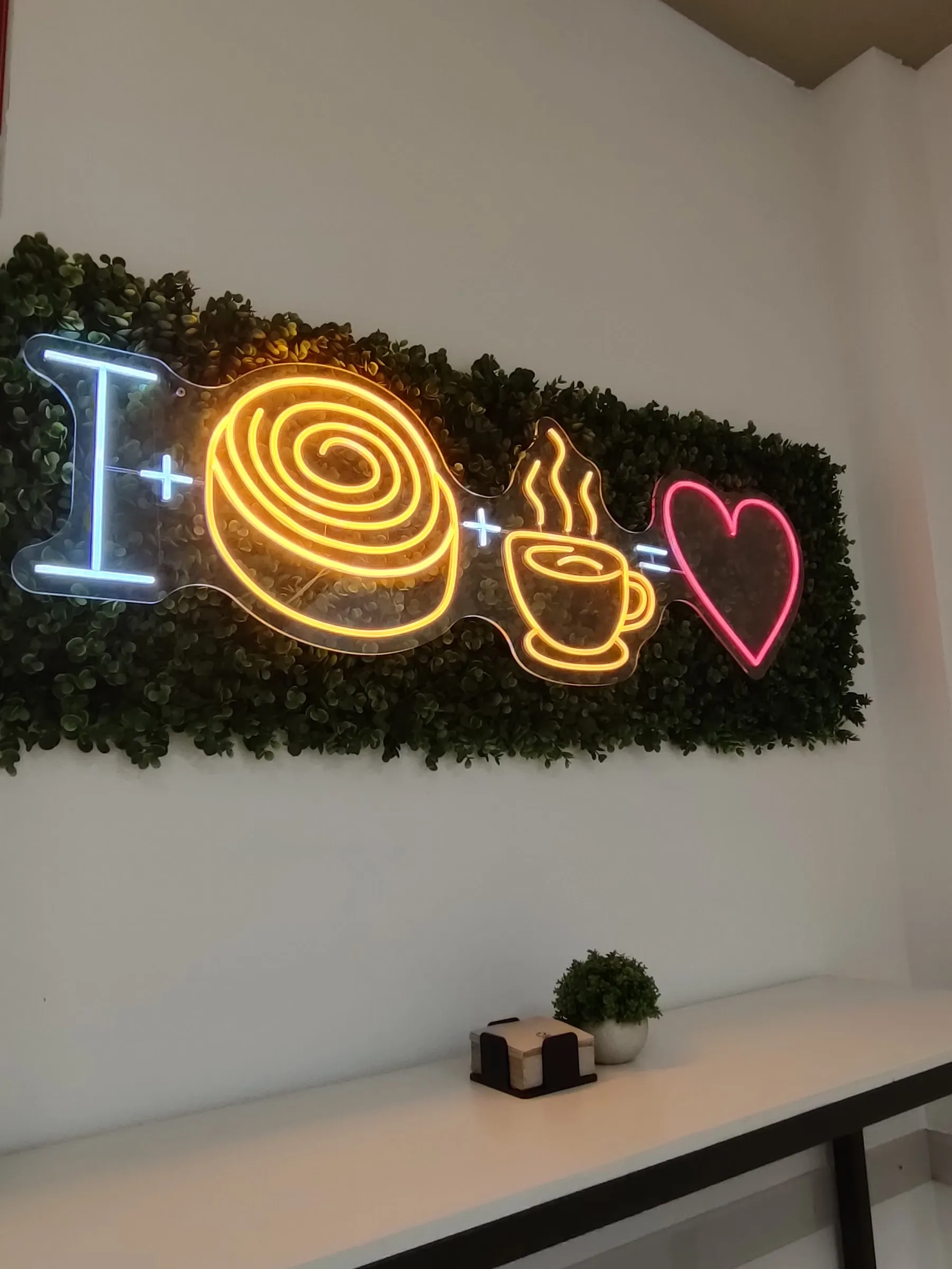 La pared del local: un neón con una I, un rollo de canela en espiral, una taza humeante y un corazón, sobre un panel verde
